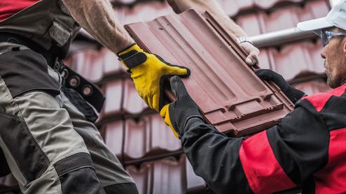 Zwei Handwerker übergeben eine Dachziegel auf einem Steildach, während eine Leiter im Hintergrund sichtbar ist.