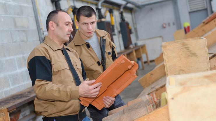 Zwei Männer in Arbeitskleidung betrachten Ziegelsteine an einem Tisch in einer Werkstatt.